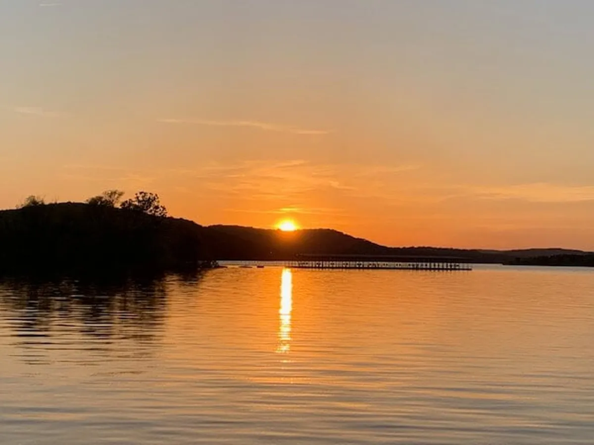 A breathtaking sunset casts warm hues over calm water, with a wooden pier stretching into Table Rock Lake and gentle hills in the background. The reflections and colors evoke a mood of ultimate relaxation.