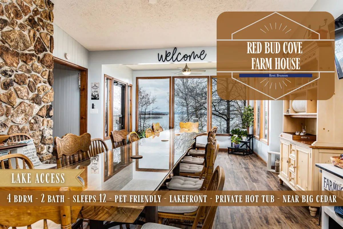 An interior dining area with a large table sits beside expansive windows overlooking the lake, with a stone wall and a welcome sign adding to the friendly ambiance. The setting is ideal for group dinners, lake views, and evening relaxation—including hot tub time.