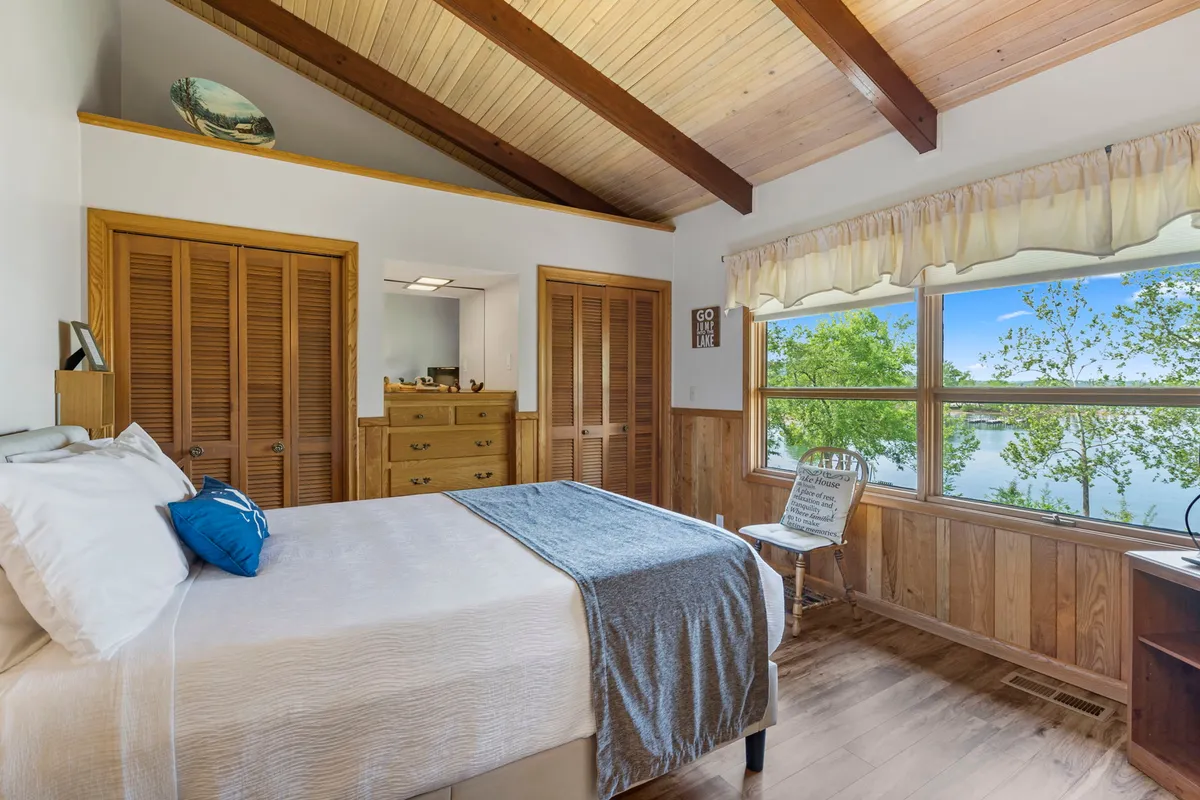 A serene bedroom featuring a comfortable bed with a blue accent pillow, large window offering scenic views, and wooden furniture that creates a tranquil, inviting ambiance. Natural light fills the space, highlighting restful touches intended for adult relaxation.