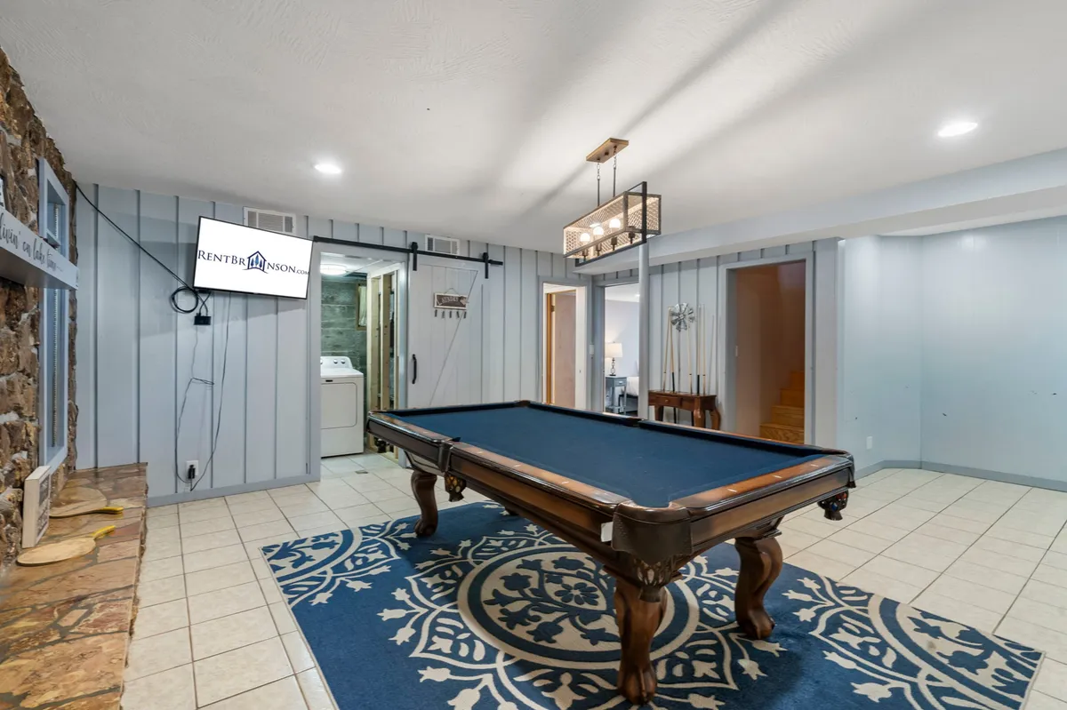 A bright and inviting recreation room with a pool table as the centerpiece, set atop a decorative rug. A stone fireplace and TV screen offer additional options for gatherings and entertainment, while a chandelier adds a touch of warmth.