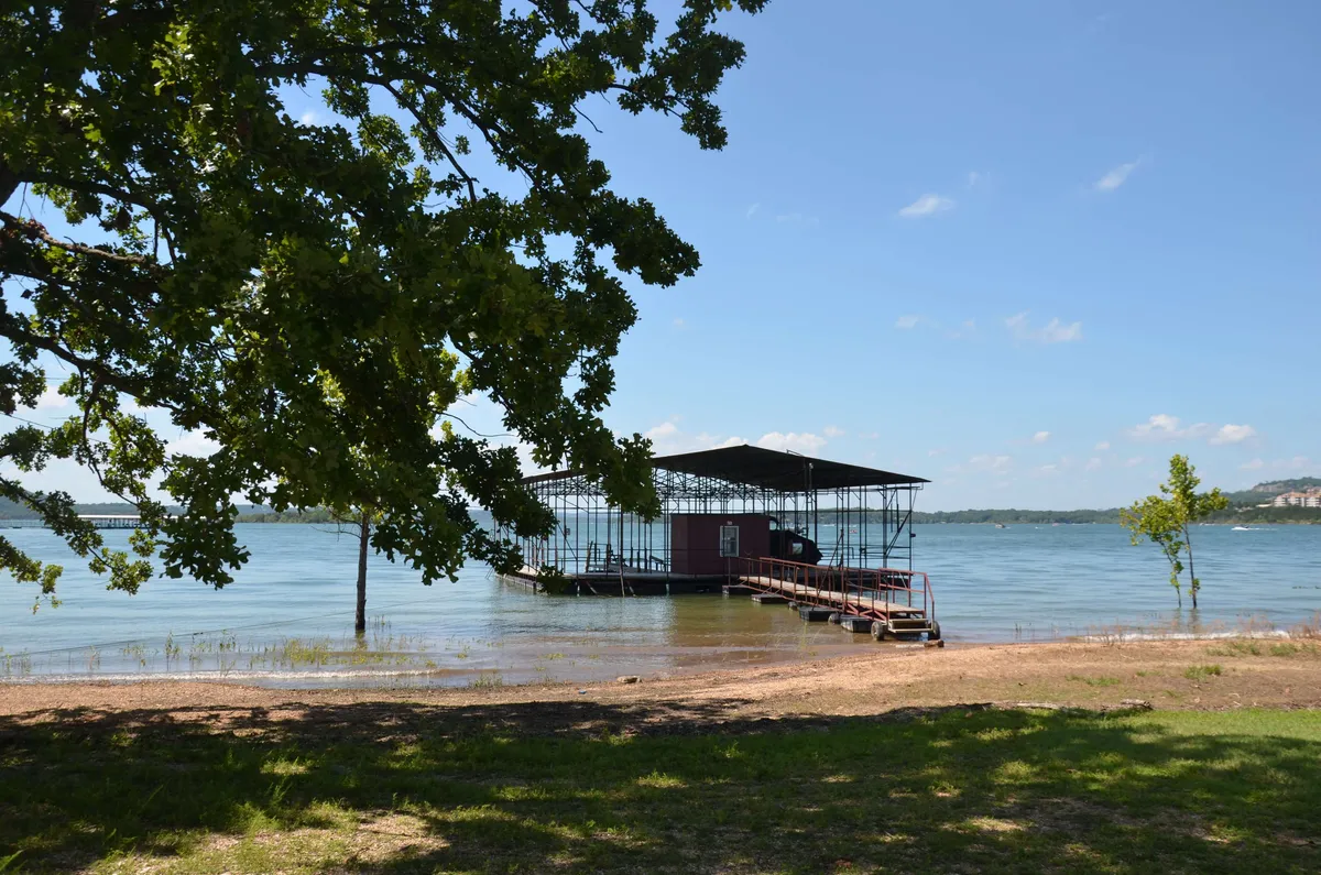 A tranquil lakeside scene featuring a dock extending over calm water, surrounded by lush greenery and a building in the background. The inviting dock is perfect for launching kayaks or paddleboards, and the water looks ideal for both paddling and fishing. The peaceful setting suggests opportunities for relaxation and water-based fun.