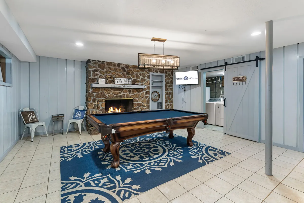 A warmly lit game room featuring a pool table, a cozy rug, a fireplace, and a TV mounted on the wall. The inviting space is set up for friendly competition and evening relaxation, with comfortable seating and a casual lounge atmosphere.