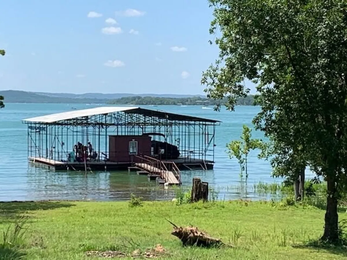 A floating dock set along the tranquil lakefront, surrounded by mature trees and with a waterfront structure nearby. The scene is peaceful yet inviting, suggesting the communal aspect of enjoying lake activities together.