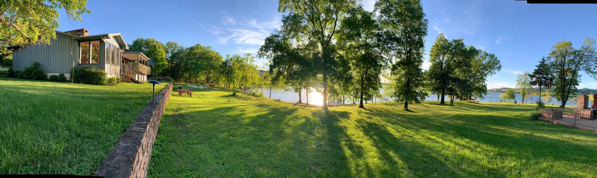 This image features a serene lake with trees surrounding a spacious house and a well-kept lawn. The outdoor setting invites activities such as picnicking, fishing, boating, and relaxing, with an emphasis on sunset views and lakeside enjoyment.