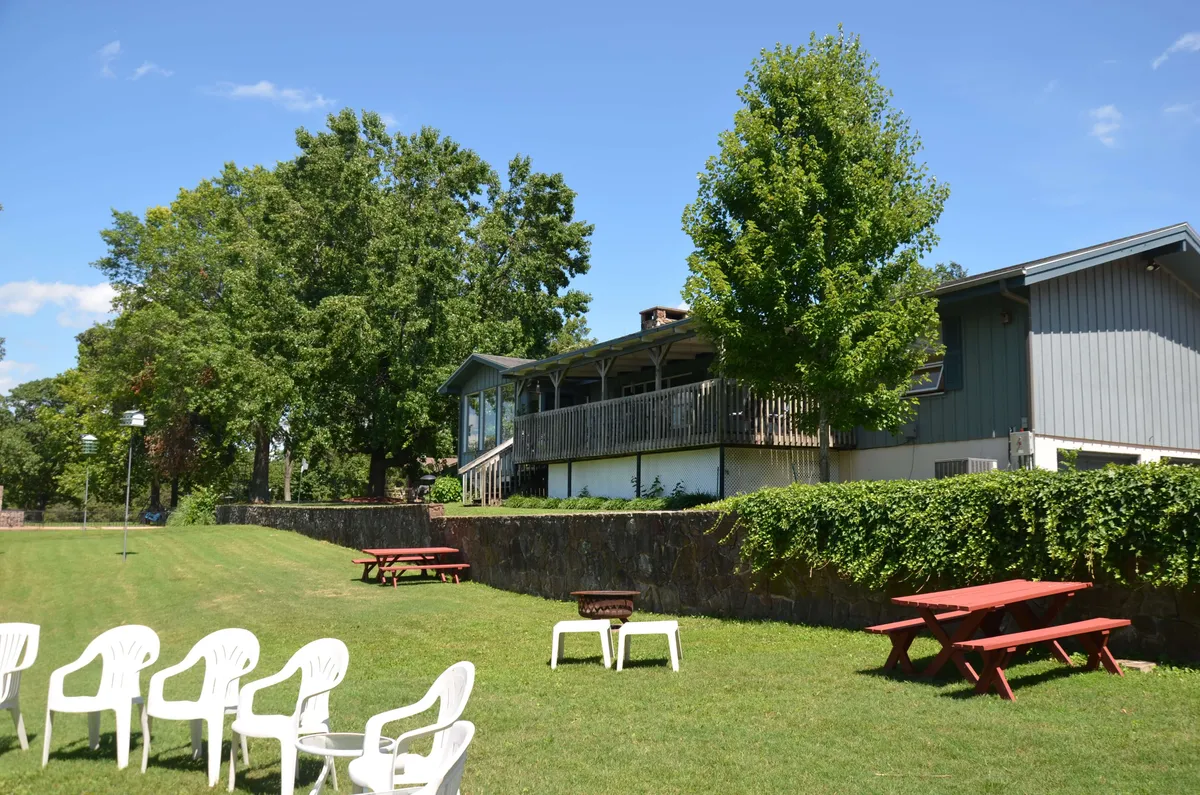 A broad green lawn with outdoor seating and a wooden structure shaded by mature trees. The space is clearly intended for group gatherings, picnics, and lawn games, surrounded by a relaxed and inviting atmosphere.