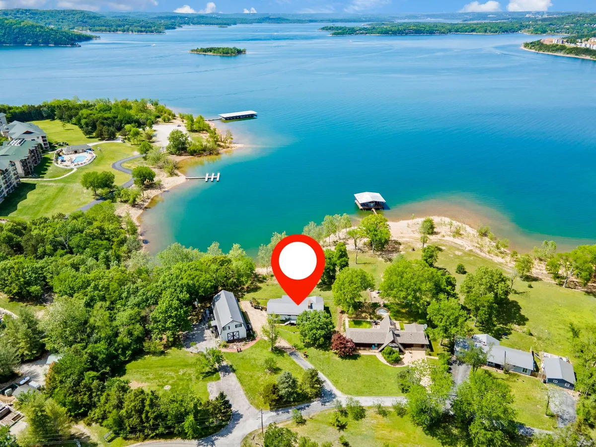 A panoramic view of Table Rock Lake with calm waters surrounded by lush greenery, several houses dotting the shoreline, and a dock extending into the lake. Boaters can be seen enjoying the water, while others picnic on the nearby grass and explore the lakeside.