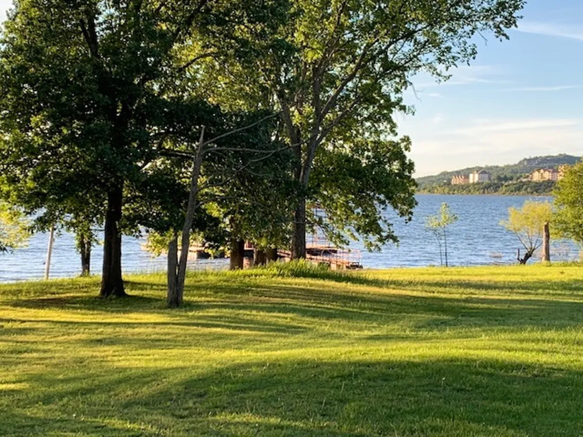 A peaceful lakeside scene with trees lining the shore and distant buildings visible across calm water. The natural setting invites picnicking, walking, and wildlife watching along the lake.