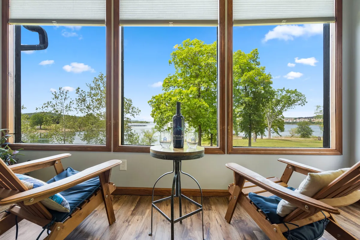 The hero image showcases a cozy lodge interior with large windows framing a breathtaking view of Table Rock Lake. Wooden chairs are arranged facing the water, with a wine bottle and glasses set on a small table—inviting guests to relax and savor the sunset. The tranquil setting emphasizes the direct lakefront access and the serene mood of the property.