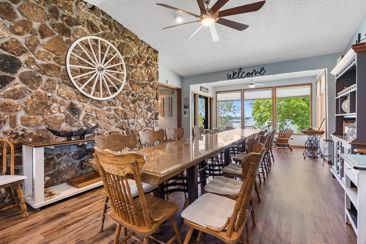 A long dining table surrounded by chairs is set against a rustic stone wall, with large windows framing a picturesque outdoor view. The space is filled with warm wood and natural light, perfect for group meals and lively conversation.