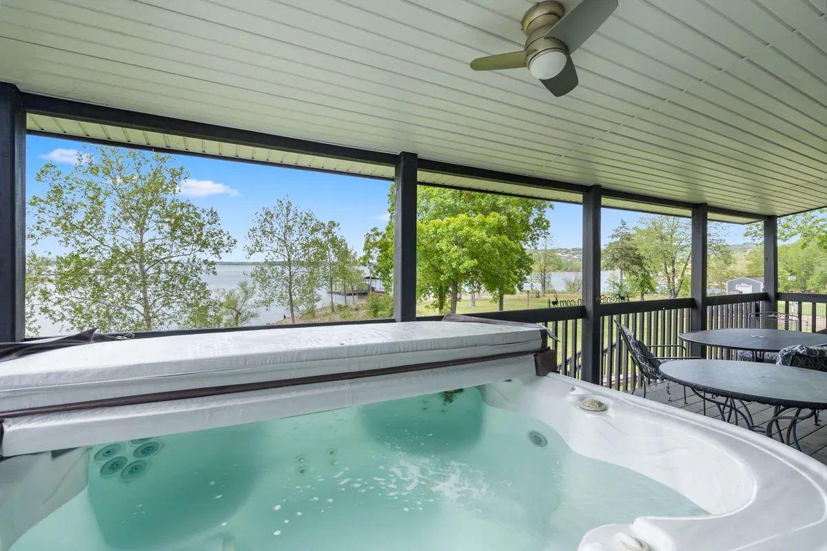 An exterior view of a covered deck featuring a private hot tub, surrounded by trees and offering tranquil views of the lake. An outdoor seating area invites guests to relax and take in the serene setting.