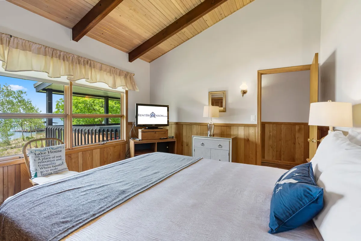 A cozy bedroom with a comfortable bed adorned with decorative pillows, large windows that bring in natural light and offer scenic views, a TV on a stand, and warm wooden paneling. The space exudes comfort and privacy, ideal for unwinding after an active day.