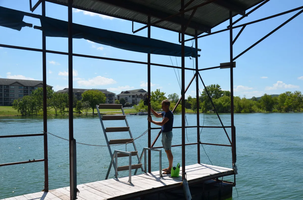 This image features a waterfront dock with a person working or fishing, calm water, and a scenic landscape in the background. The dock structure suggests a shared amenity ideal for fishing, swimming, or simply enjoying the lake views.
