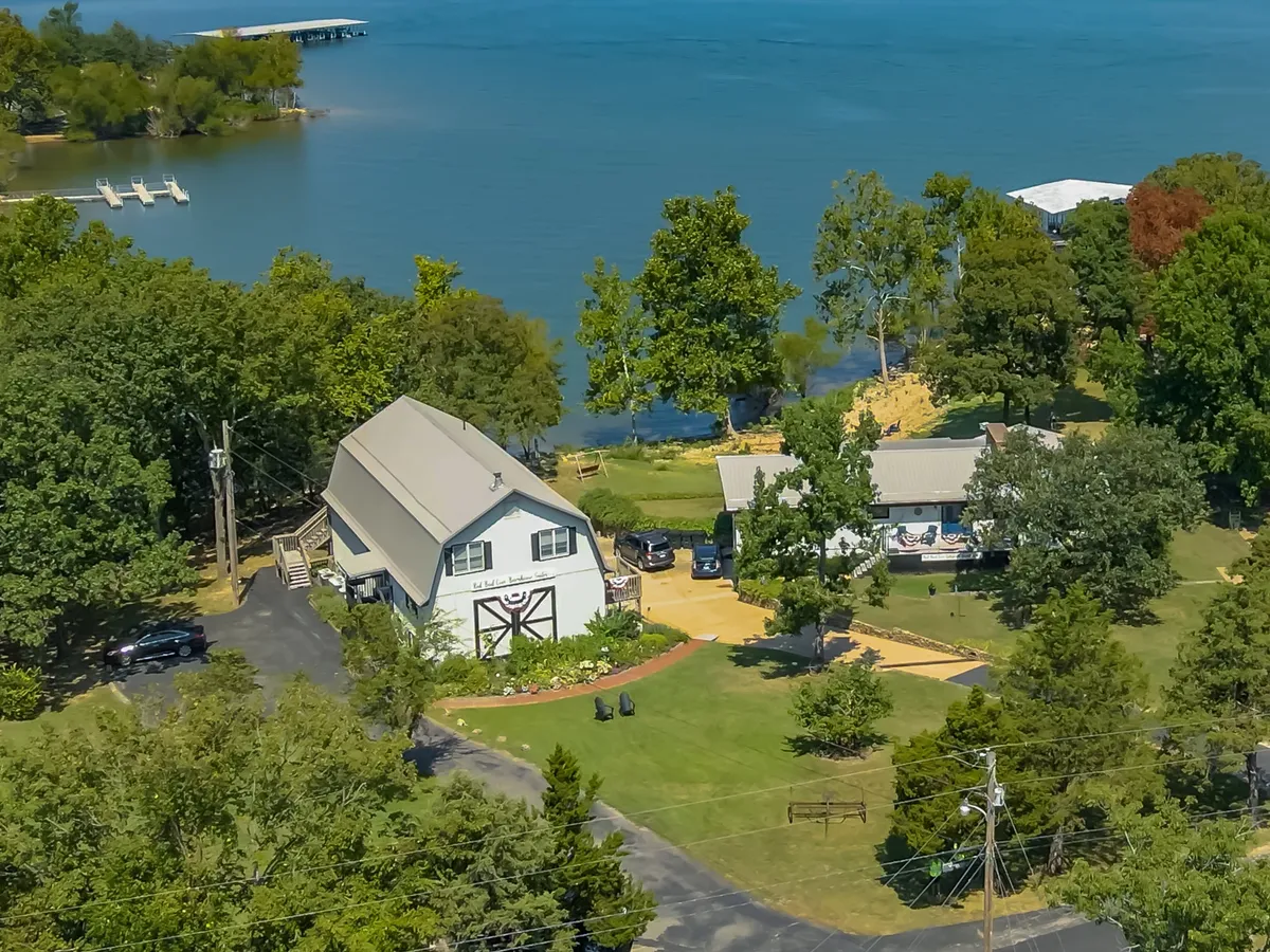 This image presents a tranquil lakefront scene with the main house set against a backdrop of calm water and surrounding trees. A patio area offers a spot to relax close to the shoreline, highlighting the property's direct water access.